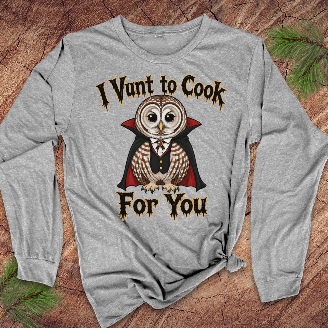 A heather gray long-sleeve t-shirt with a graphic of a vampire owl and the text 'I Vunt to Cook For You' in a gothic font, displayed flat on a rustic wood background with pine needles.