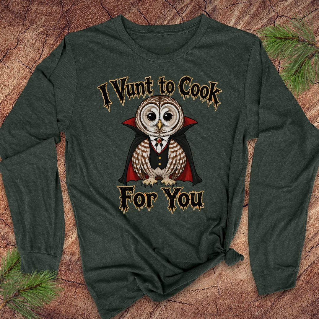 A dark forest green long-sleeve t-shirt with a graphic of a cute owl dressed as a vampire, with the text 'I Vunt to Cook For You' in a gothic font. The shirt is laid flat on a rustic wood background with pine needles in the corners.