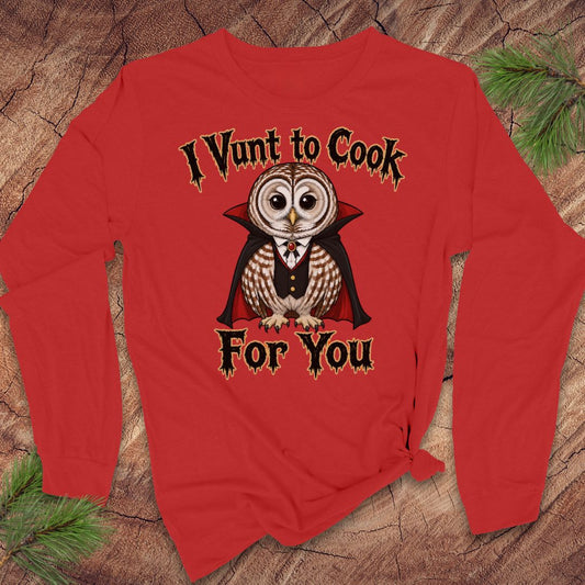A red long-sleeve t-shirt with a graphic of a vampire owl and the text 'I Vunt to Cook For You' in a stylized font, displayed flat on a rustic wood background with pine branches.