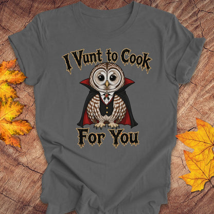 A dark gray graphic t-shirt featuring a cartoon vampire owl with the text 'I Vunt to Cook For You' in a gothic font. The t-shirt is displayed flat on a rustic wood background decorated with autumn leaves.