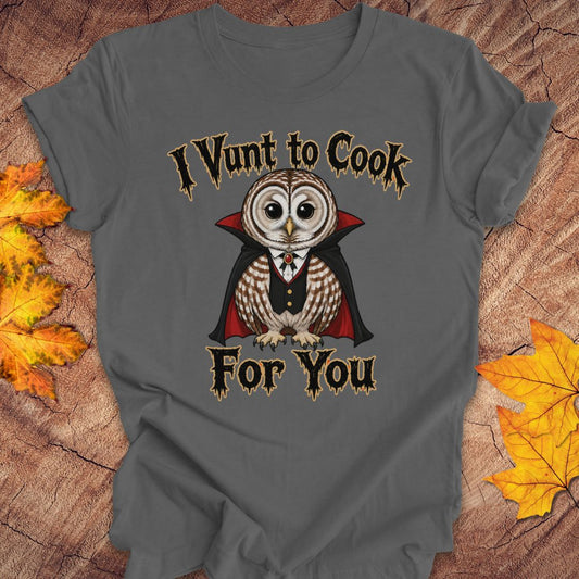 A dark gray graphic t-shirt featuring a cartoon vampire owl with the text 'I Vunt to Cook For You' in a gothic font. The t-shirt is displayed flat on a rustic wood background decorated with autumn leaves.