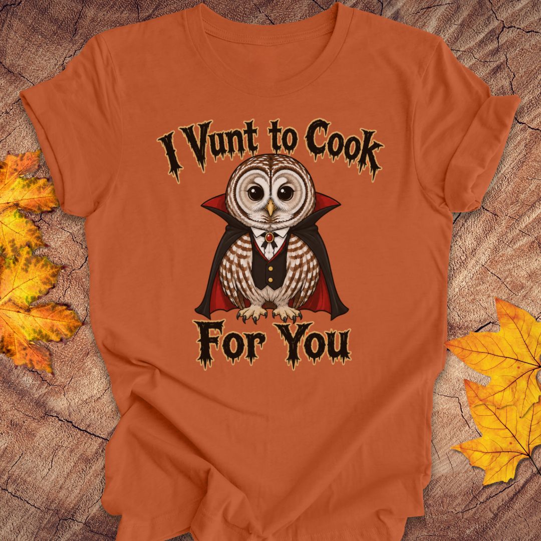 An orange short-sleeve graphic T-shirt laid flat on a rustic wood surface with autumn leaves. The shirt features a vampire owl illustration with the text 'I Vunt to Cook For You' in a stylized font.