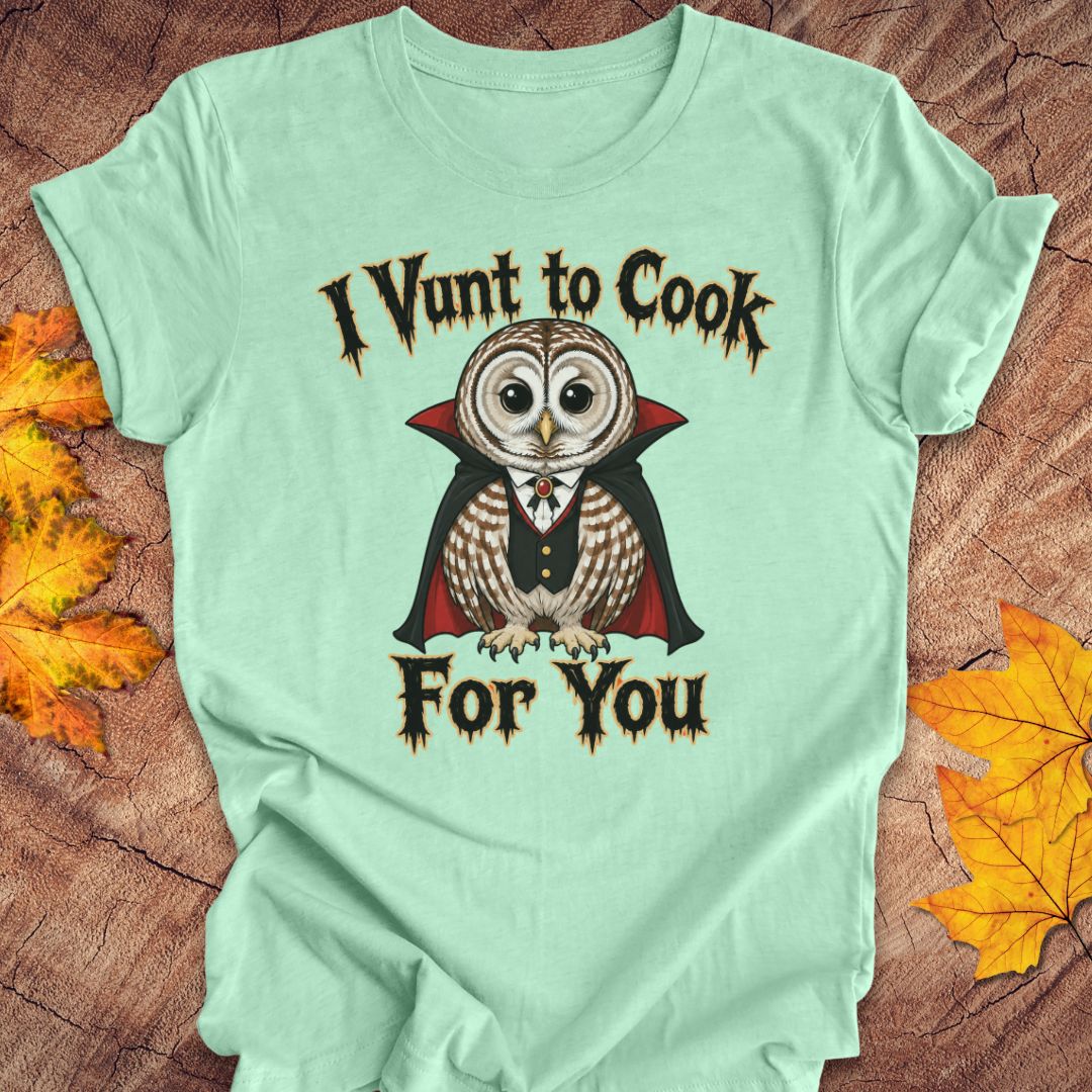 A mint green graphic t-shirt laid flat on a rustic wood background with autumn leaves. The shirt features a vampire owl illustration with the text 'I Vunt to Cook For You' in a gothic font.