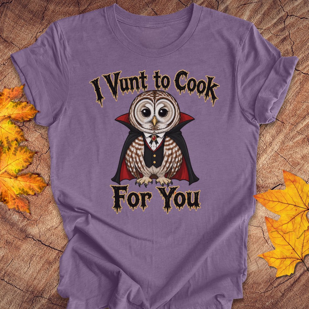 A purple short-sleeve graphic T-shirt laid flat on a wood surface with autumn leaves around it. The shirt features a vampire owl illustration with the text 'I Vunt to Cook For You' in a gothic font.