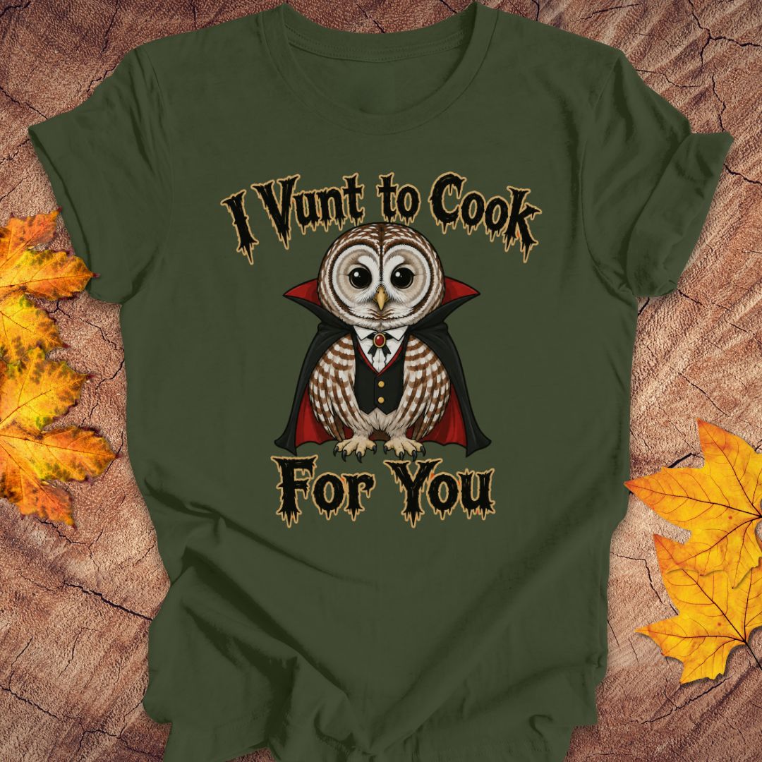 An olive green short-sleeve t-shirt laid flat on a rustic wood background with autumn leaves. The shirt features a graphic of a cute owl dressed as a vampire with the text 'I Vunt to Cook For You' in a spooky font.