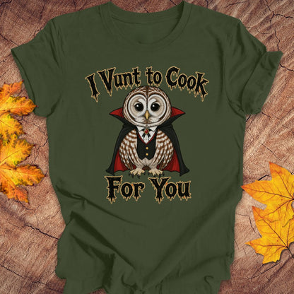 An olive green short-sleeve t-shirt laid flat on a rustic wood background with autumn leaves. The shirt features a graphic of a cute owl dressed as a vampire with the text 'I Vunt to Cook For You' in a spooky font.