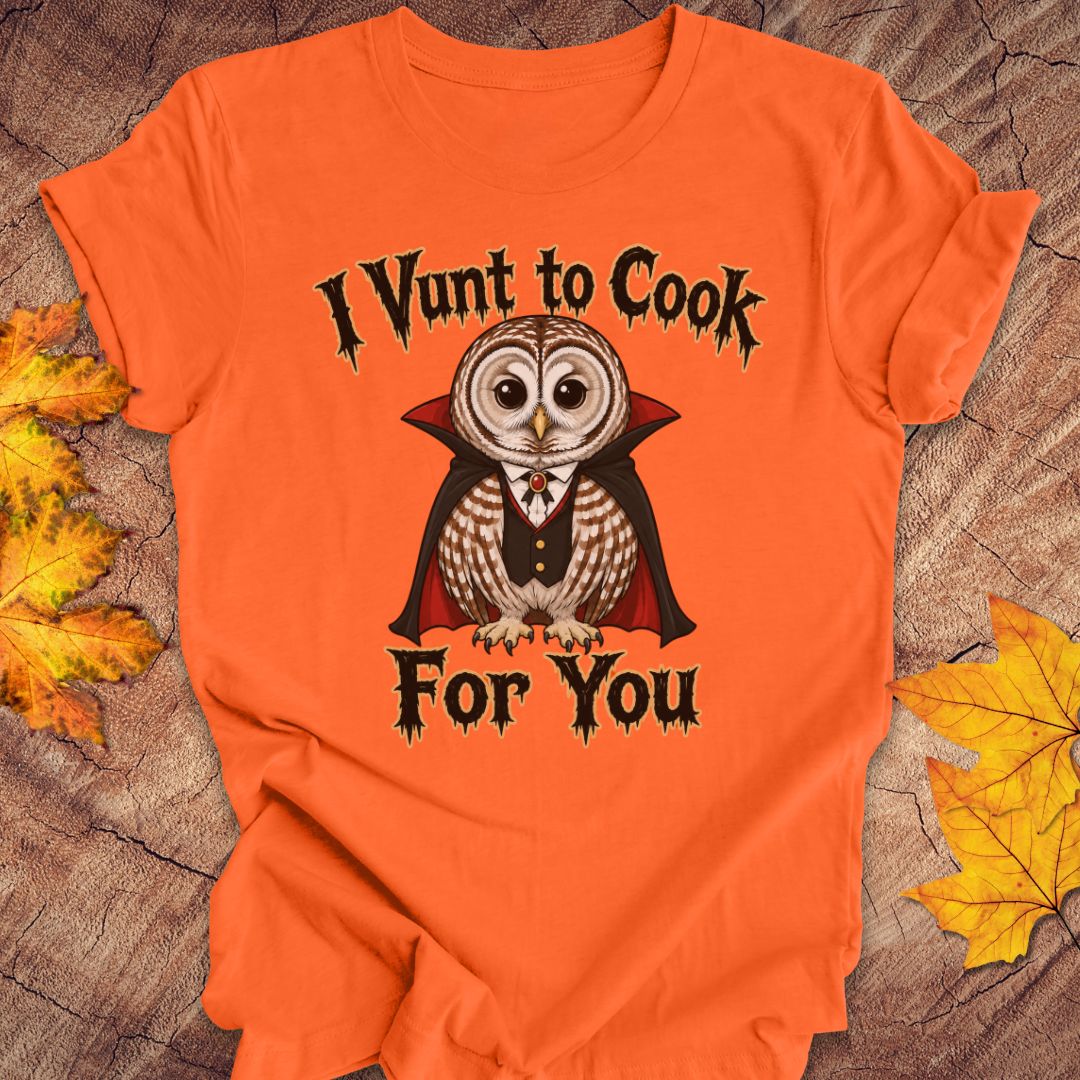 An orange short-sleeve t-shirt laid flat on a rustic wood surface with autumn leaves. The shirt features a graphic of a vampire owl wearing a cape and bow tie, with the text 'I Vunt to Cook For You' in a gothic font.