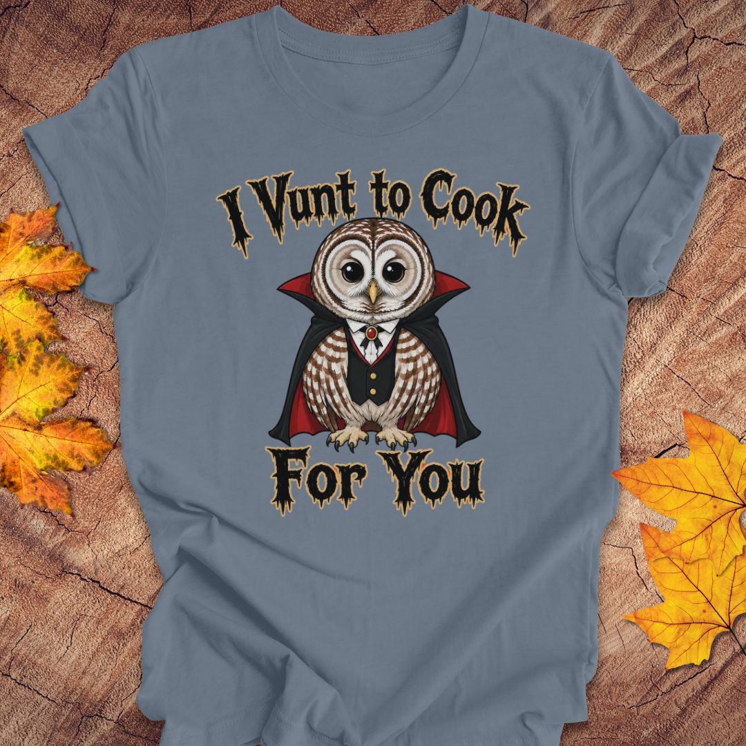 A slate blue graphic t-shirt laid flat on a rustic wood background with autumn leaves. The shirt features a graphic of a cute owl dressed as a vampire with the text 'I Vunt to Cook For You' in a gothic font.