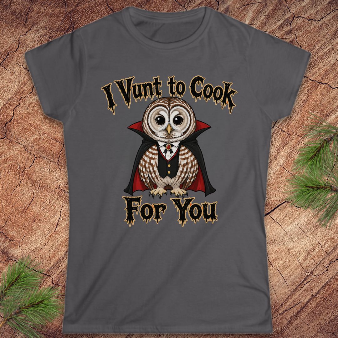 A dark gray graphic t-shirt featuring a cartoon owl dressed as a vampire with the text 'I Vunt to Cook For You' in a gothic font, displayed flat on a rustic wood background with pine needles.