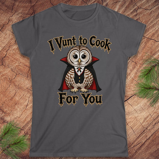 A dark gray graphic t-shirt featuring a cartoon owl dressed as a vampire with the text 'I Vunt to Cook For You' in a gothic font, displayed flat on a rustic wood background with pine needles.