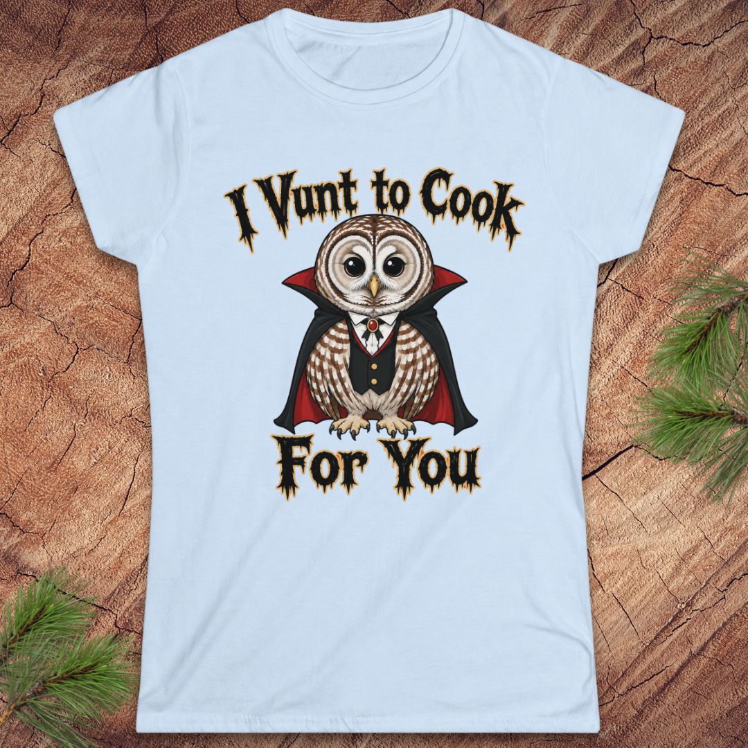 A light blue short-sleeve crewneck t-shirt featuring a graphic of a vampire owl with the text 'I Vunt to Cook For You' in a gothic font, displayed flat on a rustic wood background with pine needles.