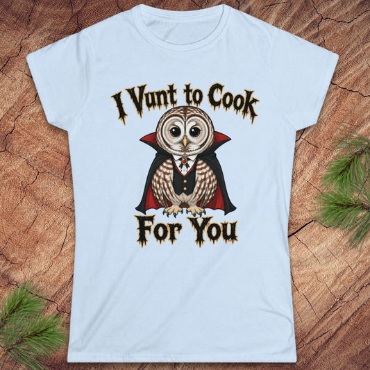A light blue short-sleeve crewneck t-shirt featuring a graphic of a vampire owl with the text 'I Vunt to Cook For You' in a gothic font, displayed flat on a rustic wood background with pine needles.