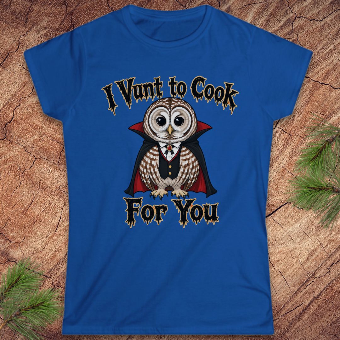 A royal blue women's short-sleeve t-shirt with a graphic of a vampire owl and the text 'I Vunt to Cook For You' in a gothic font, displayed flat on a rustic wood background with pine needles.