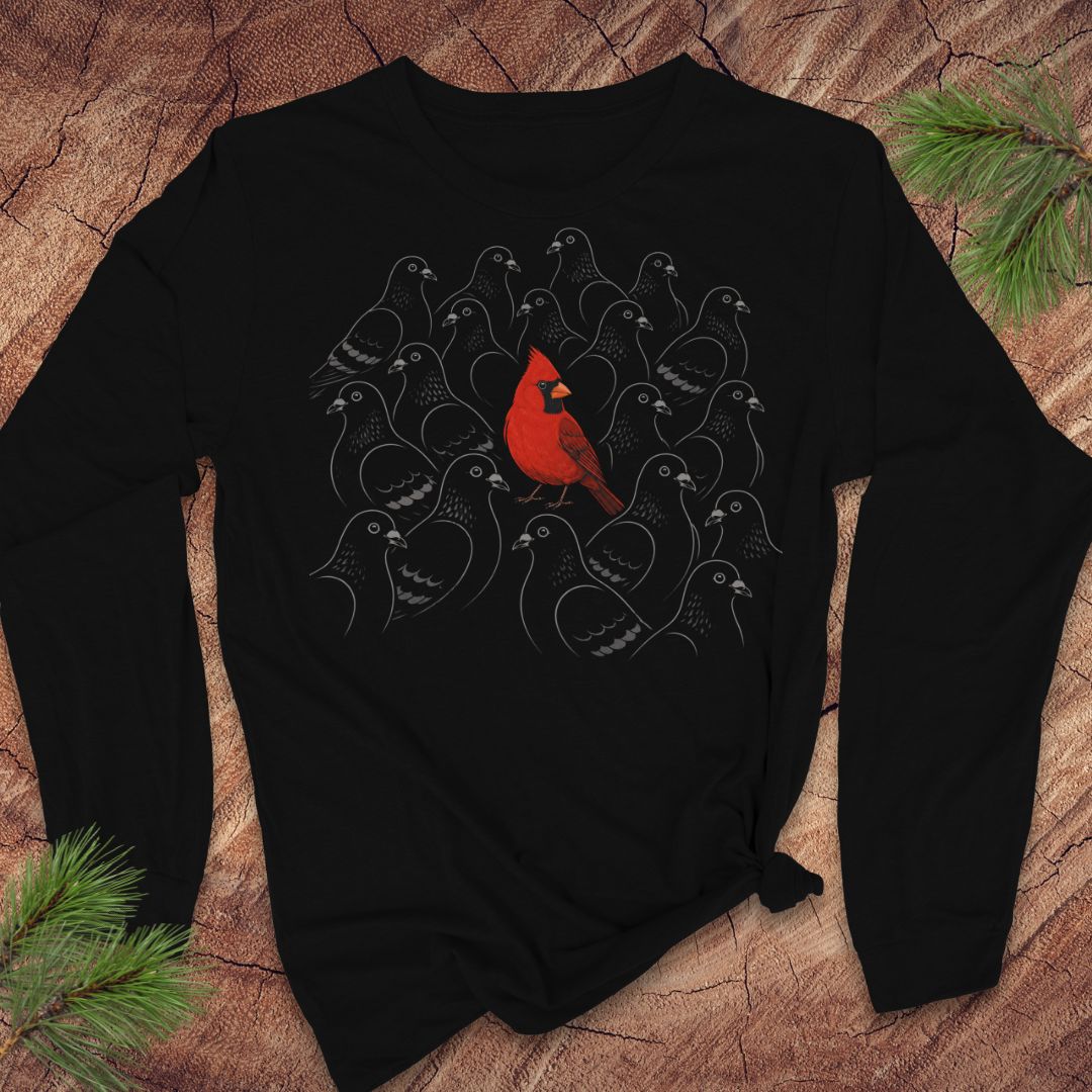 Black long-sleeve t-shirt with a bright red Cardinal standing out in a crowd of gray line-drawn pigeons, laid flat on a rustic wood background with pine branches.