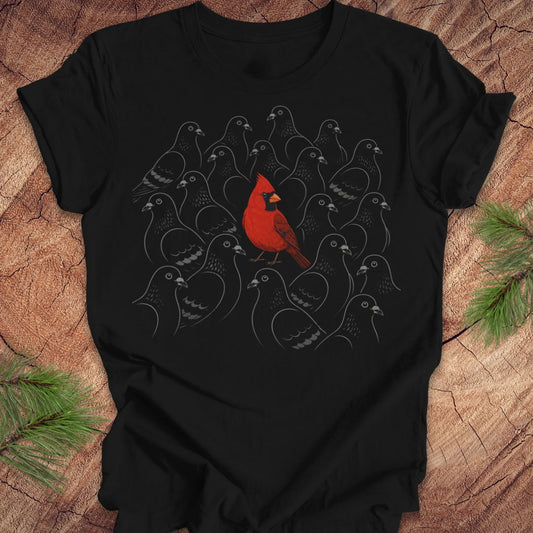 A black short-sleeve t-shirt laid flat on a rustic wood surface, featuring a graphic of a bright red cardinal perched in front of a cluster of gray bird silhouettes. Green pine branches are placed in the corners of the frame.