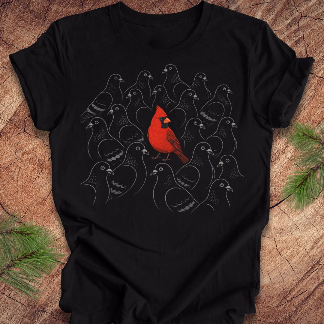 A black short-sleeve crewneck t-shirt laid flat on a rustic wood surface, featuring a graphic print of a bright red cardinal surrounded by numerous gray bird silhouettes. Pine needles are visible in the corners as styling props.