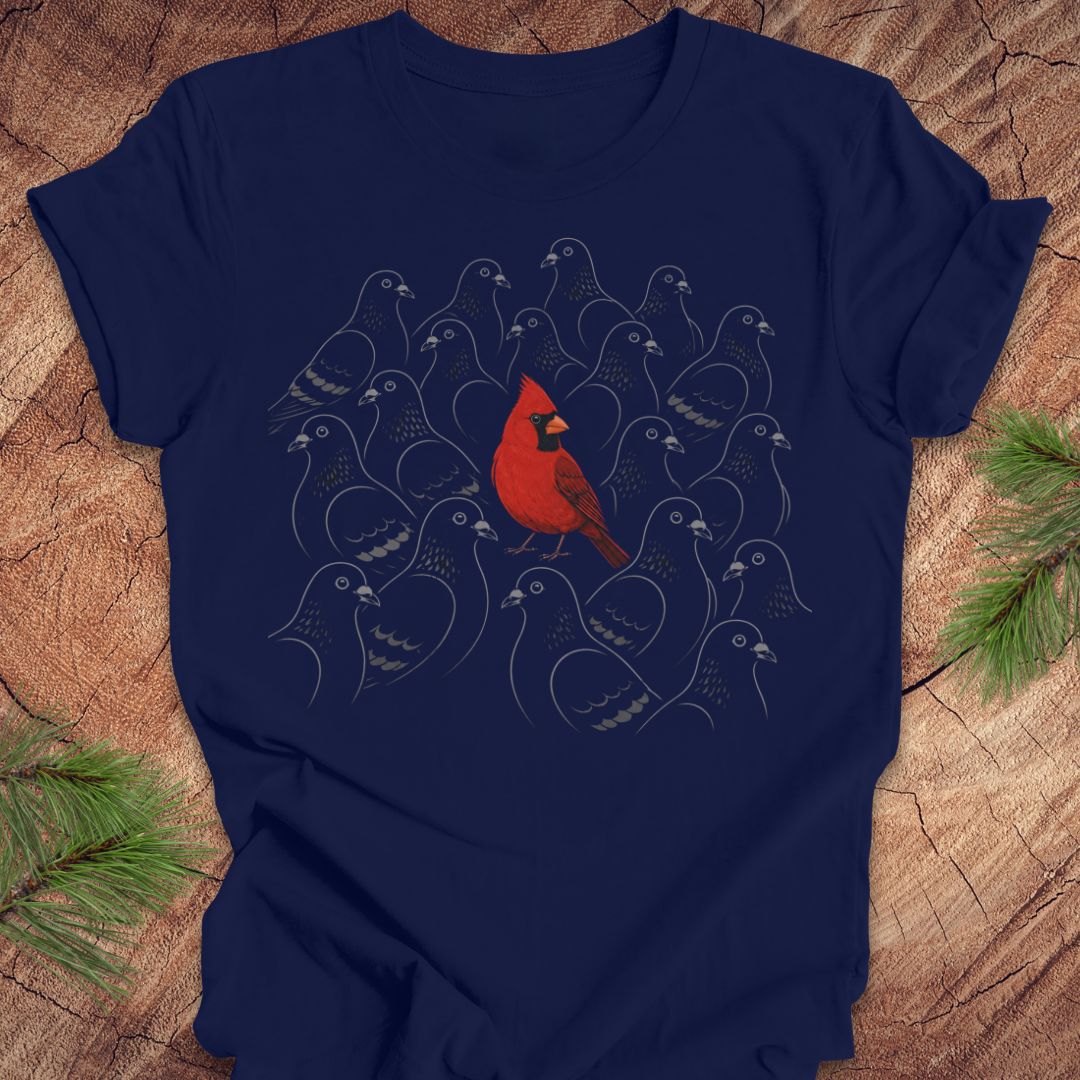 A navy blue short-sleeve t-shirt with a graphic print of a red cardinal bird surrounded by stylized gray birds, laid flat on a rustic wood background with pine branches in the corners.