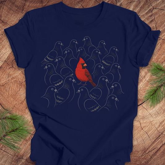 A navy blue short-sleeve t-shirt with a graphic print of a red cardinal bird surrounded by stylized gray birds, laid flat on a rustic wood background with pine branches in the corners.