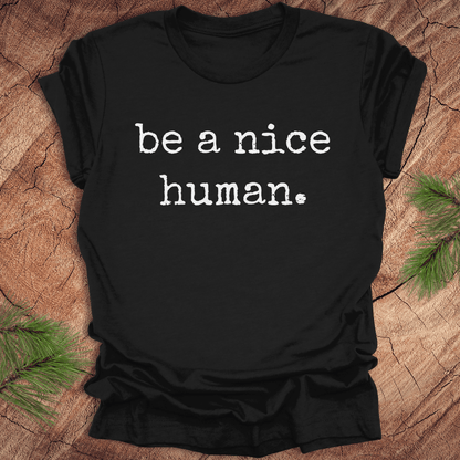 Black t-shirt with 'be a nice human.' text on a wooden surface with greenery.