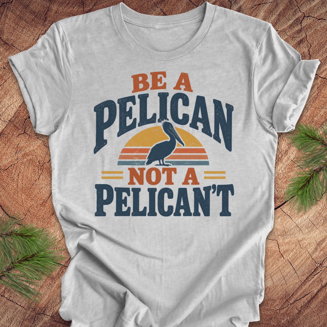Athletic heather gray t-shirt with a Brown Pelican silhouette on a retro striped sunset and the words BE A PELICAN NOT A PELICAN'T, laid flat on a rustic wood background with pine branches.