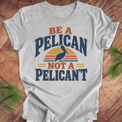 Athletic heather gray t-shirt with a Brown Pelican silhouette on a retro striped sunset and the words BE A PELICAN NOT A PELICAN'T, laid flat on a rustic wood background with pine branches.