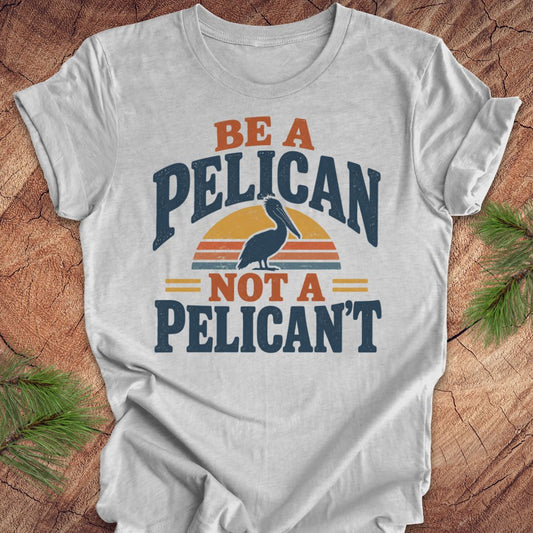 Athletic heather gray t-shirt with a Brown Pelican silhouette on a retro striped sunset and the words BE A PELICAN NOT A PELICAN'T, laid flat on a rustic wood background with pine branches.