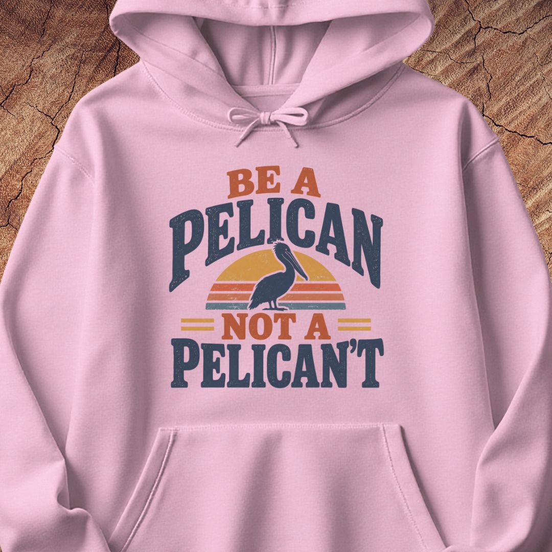 Light pink hoodie with a Brown Pelican silhouette on a retro striped sunset and the words BE A PELICAN NOT A PELICAN'T, laid flat on a rustic wood background.