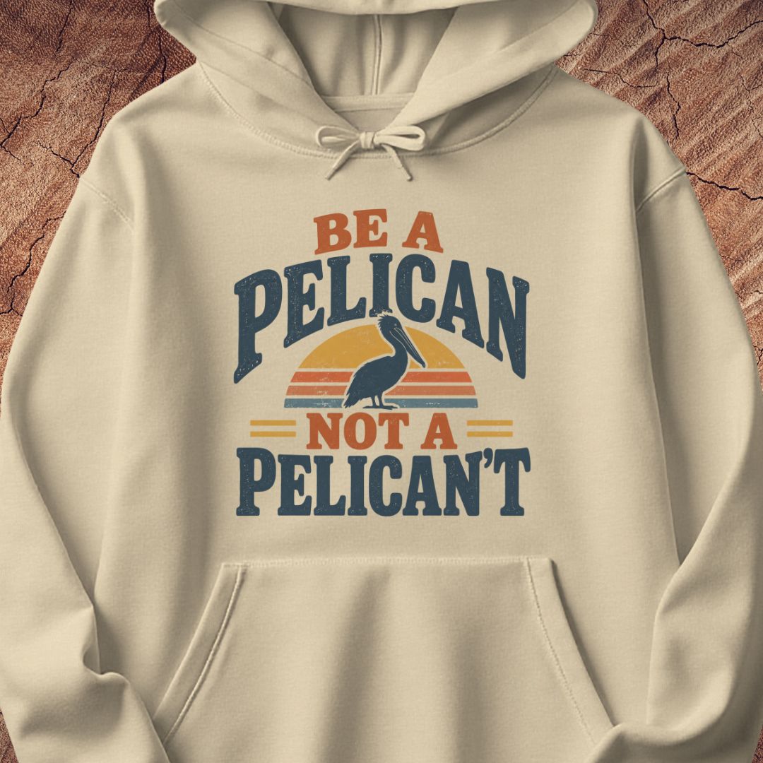 Sand beige hoodie with a Brown Pelican silhouette on a retro striped sunset and the words BE A PELICAN NOT A PELICAN'T, laid flat on a rustic wood background.