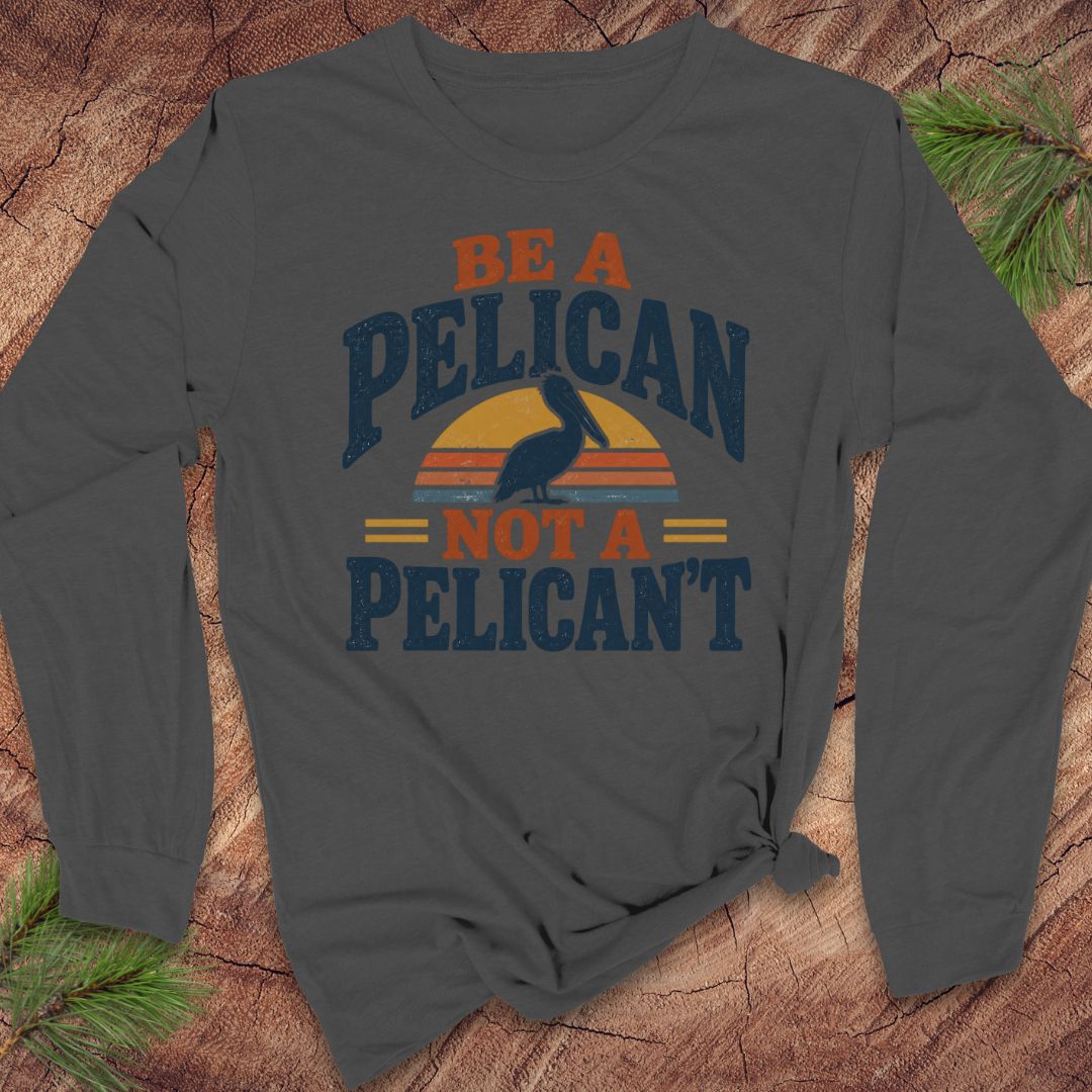 Asphalt gray long-sleeve t-shirt with a Brown Pelican silhouette on a retro striped sunset and the words BE A PELICAN NOT A PELICAN'T, laid flat on a rustic wood background with pine branches.