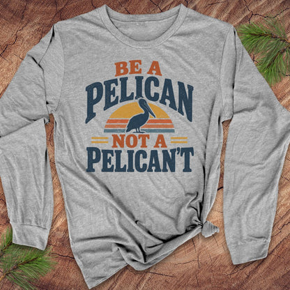 Athletic heather gray long-sleeve t-shirt with a Brown Pelican silhouette on a retro striped sunset and the words BE A PELICAN NOT A PELICAN'T, laid flat on a rustic wood background with pine branches.