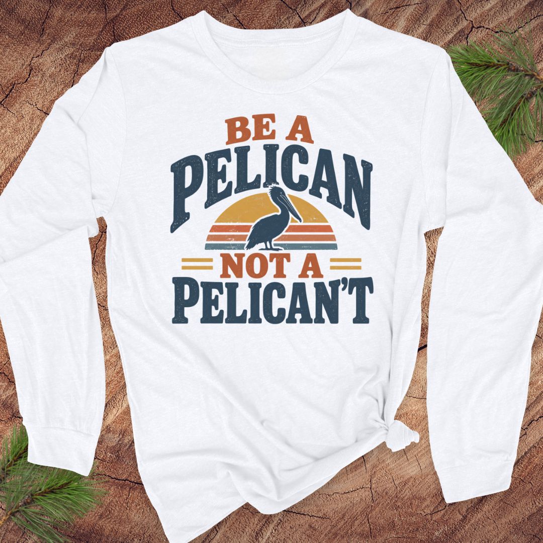 White long-sleeve t-shirt with a Brown Pelican silhouette on a retro striped sunset and the words BE A PELICAN NOT A PELICAN'T, laid flat on a rustic wood background with pine branches.