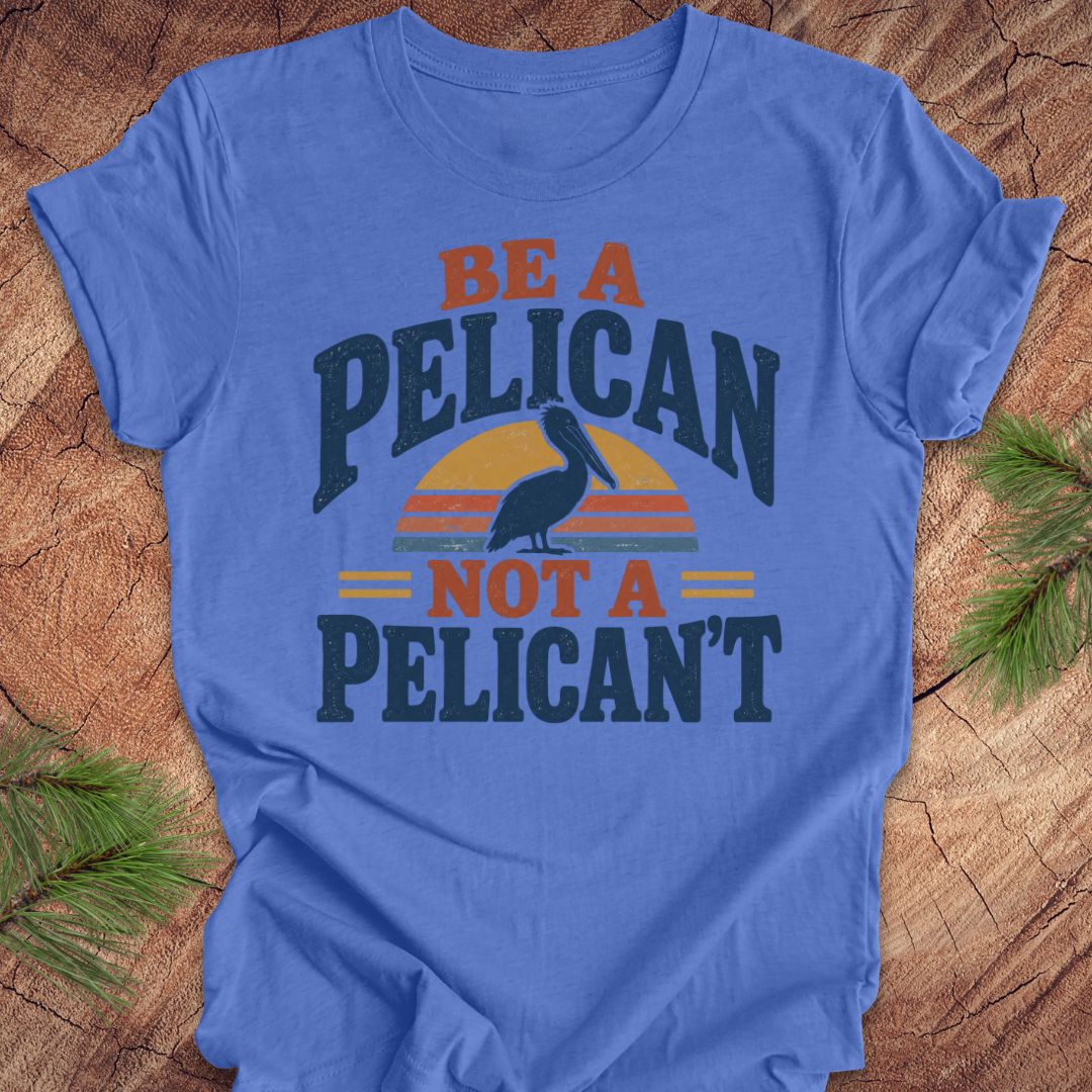 Heather Columbia blue t-shirt with a Brown Pelican silhouette on a retro striped sunset and the words BE A PELICAN NOT A PELICAN'T, laid flat on a rustic wood background with pine branches.