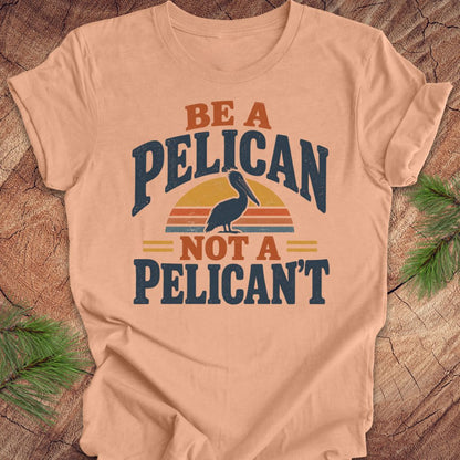 Heather peach t-shirt with a Brown Pelican silhouette on a retro striped sunset and the words BE A PELICAN NOT A PELICAN'T, laid flat on a rustic wood background with pine branches.