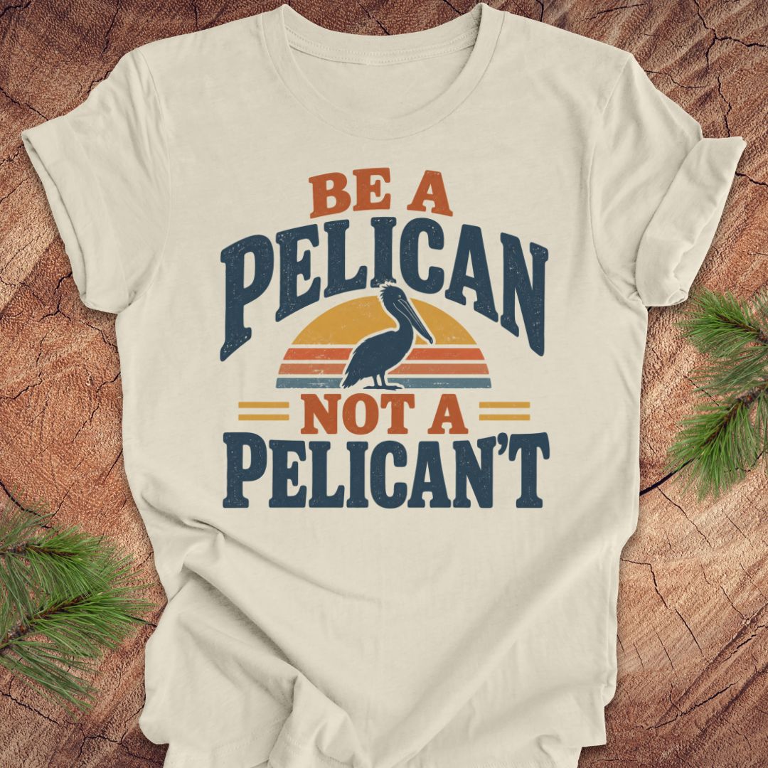 Natural cream t-shirt with a Brown Pelican silhouette on a retro striped sunset and the words BE A PELICAN NOT A PELICAN'T, laid flat on a rustic wood background with pine branches.