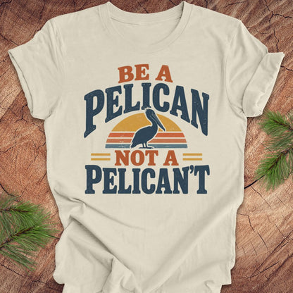 Natural cream t-shirt with a Brown Pelican silhouette on a retro striped sunset and the words BE A PELICAN NOT A PELICAN'T, laid flat on a rustic wood background with pine branches.