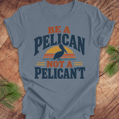 Steel blue t-shirt with a Brown Pelican silhouette on a retro striped sunset and the words BE A PELICAN NOT A PELICAN'T, laid flat on a rustic wood background with pine branches.