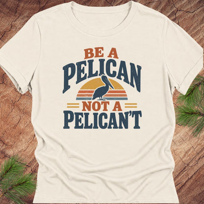 Natural cream women's relaxed t-shirt with a Brown Pelican silhouette on a retro striped sunset and the words BE A PELICAN NOT A PELICAN'T, laid flat on a rustic wood background with pine branches.