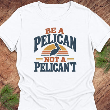 White women's relaxed t-shirt with a Brown Pelican silhouette on a retro striped sunset and the words BE A PELICAN NOT A PELICAN'T, laid flat on a rustic wood background with pine branches.
