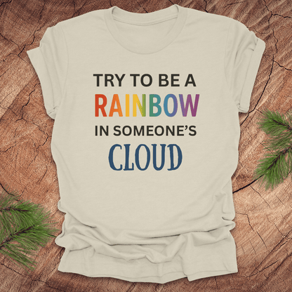 Beige t-shirt with colorful text 'Try to be a rainbow in someone's cloud' on a wooden surface