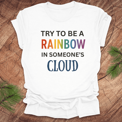 White t-shirt with colorful text 'Try to be a rainbow in someone's cloud' on a wooden surface