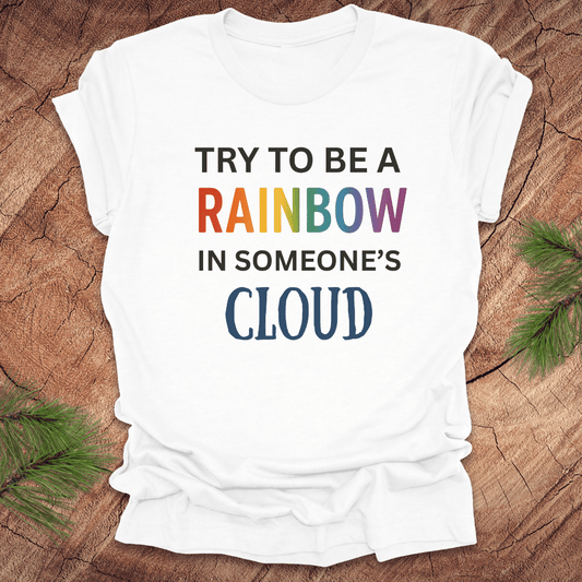 White t-shirt with colorful text 'Try to be a rainbow in someone's cloud' on a wooden surface