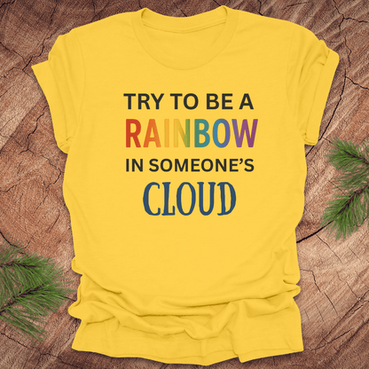 Yellow t-shirt with colorful text 'Try to be a rainbow in someone's cloud' on a wooden surface