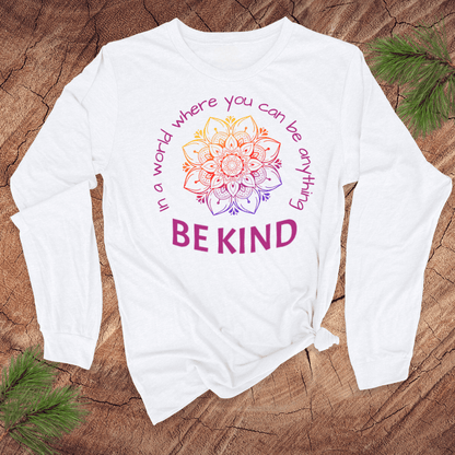 White long-sleeve shirt with a colorful mandala and 'Be Kind' text on a wooden surface.