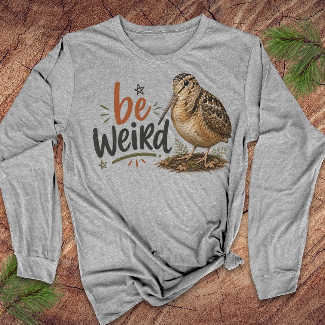 A heather gray long-sleeve t-shirt with a graphic print of a brown woodcock bird and the text 'be Weird' in orange, black, and green, laid flat on a rustic wood background with pine needles.