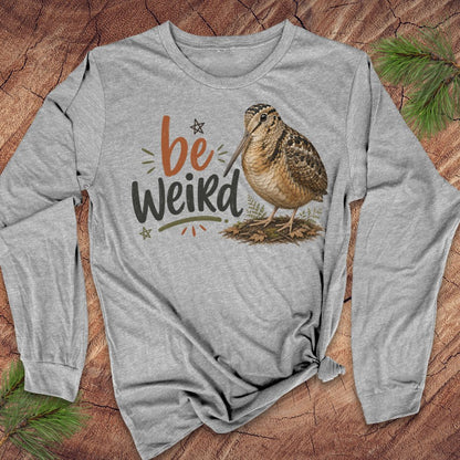 A heather gray long-sleeve t-shirt with a graphic print of a brown woodcock bird and the text 'be Weird' in orange, black, and green, laid flat on a rustic wood background with pine needles.