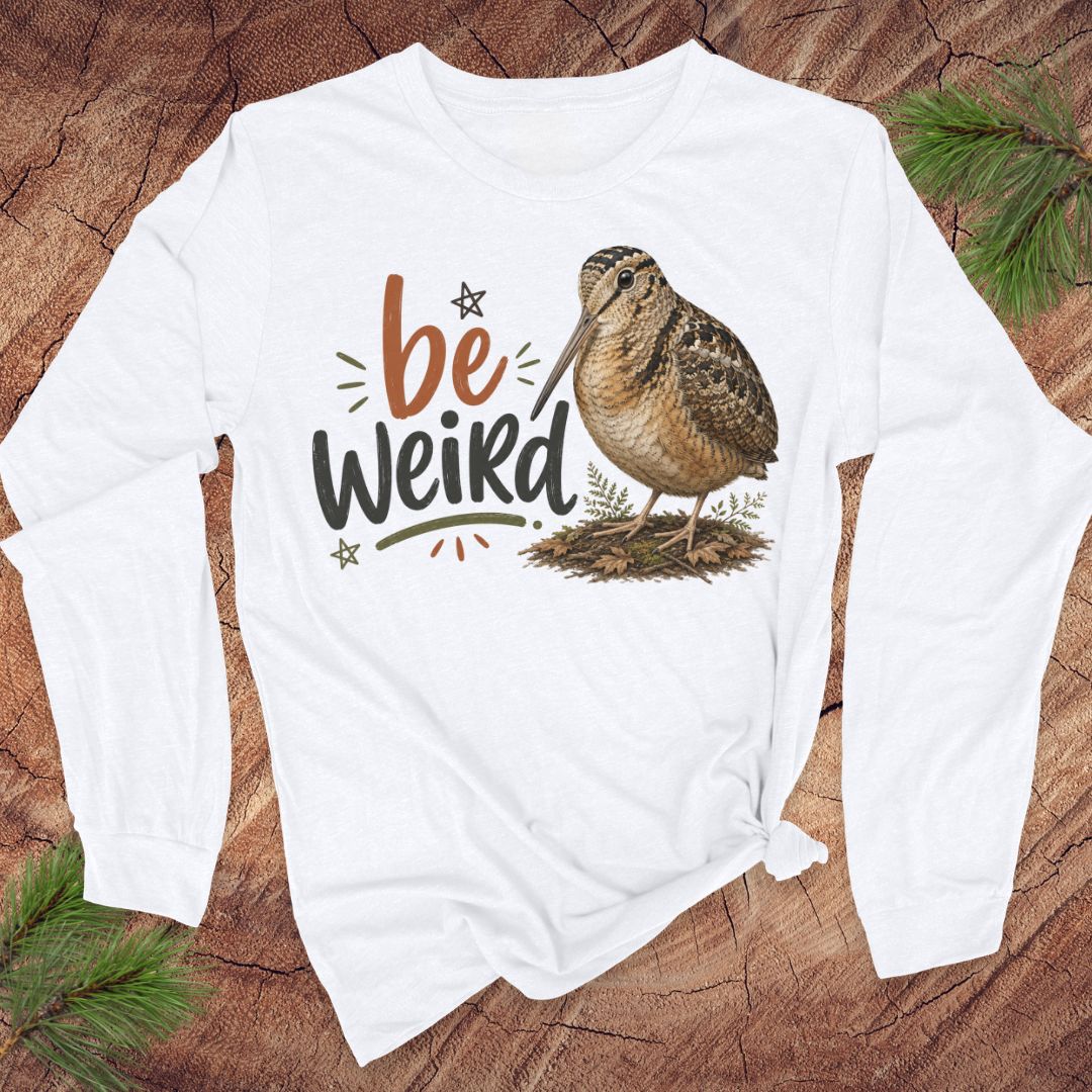 A white long-sleeve t-shirt with a graphic print of a brown woodcock bird and the text 'be Weird' in orange and black, laid flat on a rustic wood background with pine branches.