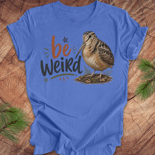 A blue graphic t-shirt featuring the text 'be Weird' in orange and dark gray, alongside an illustration of an american woodcock bird. The t-shirt is displayed flat on a rustic wood background decorated with pine branches.