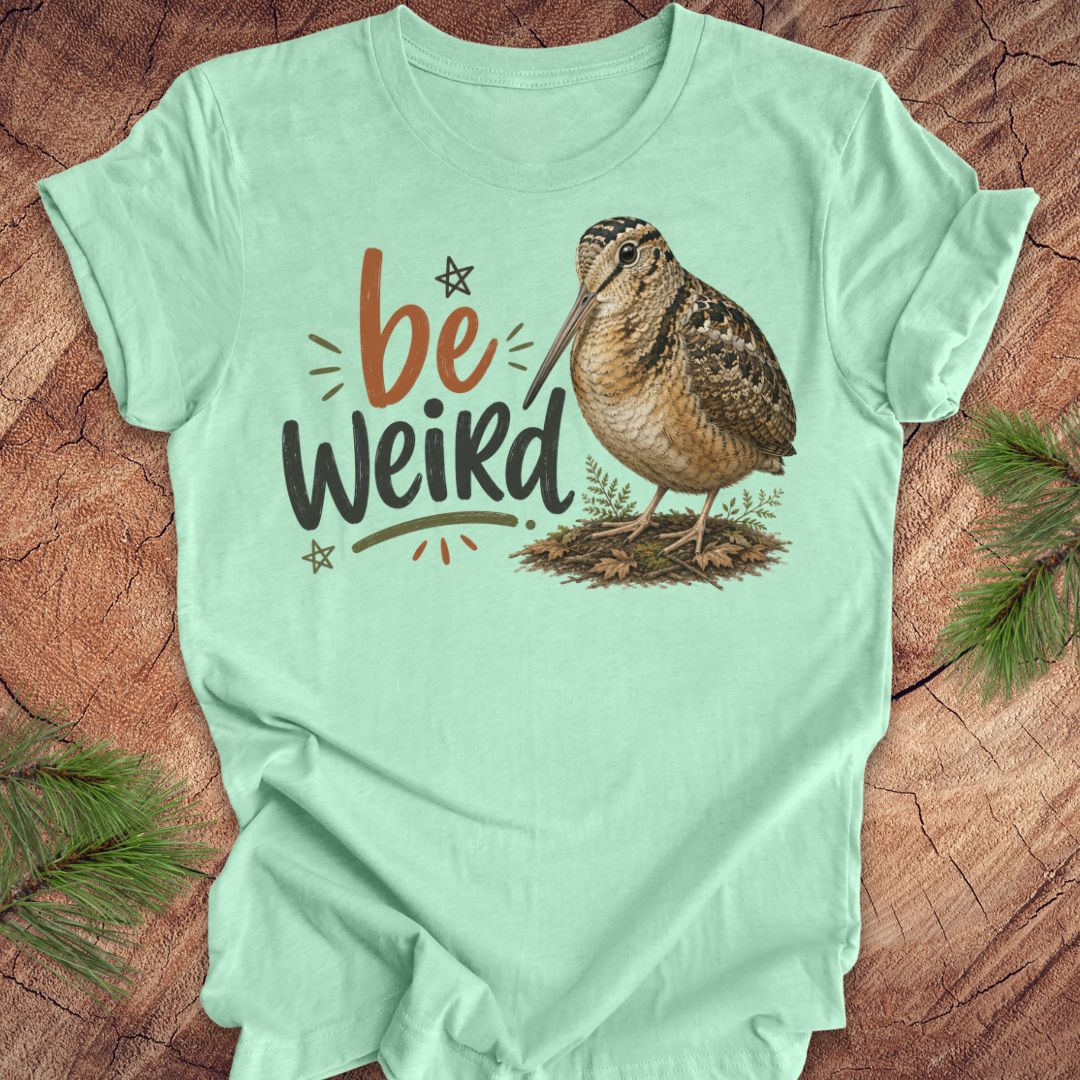 A mint green graphic t-shirt featuring the text 'be Weird' in orange and dark gray, alongside an illustration of an american woodcock bird. The t-shirt is displayed flat on a rustic wood background decorated with pine branches.