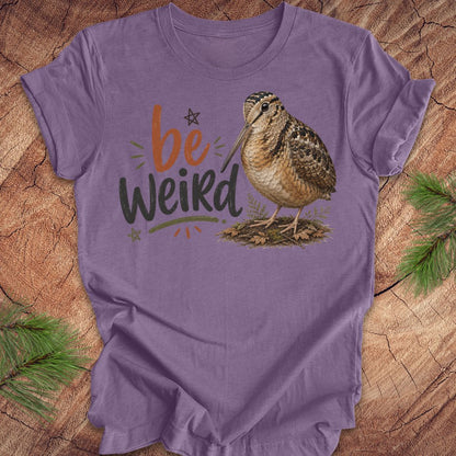 A purple graphic t-shirt featuring the text 'be Weird' in orange and dark gray, alongside an illustration of an american woodcock bird. The t-shirt is displayed flat on a rustic wood background decorated with pine branches.