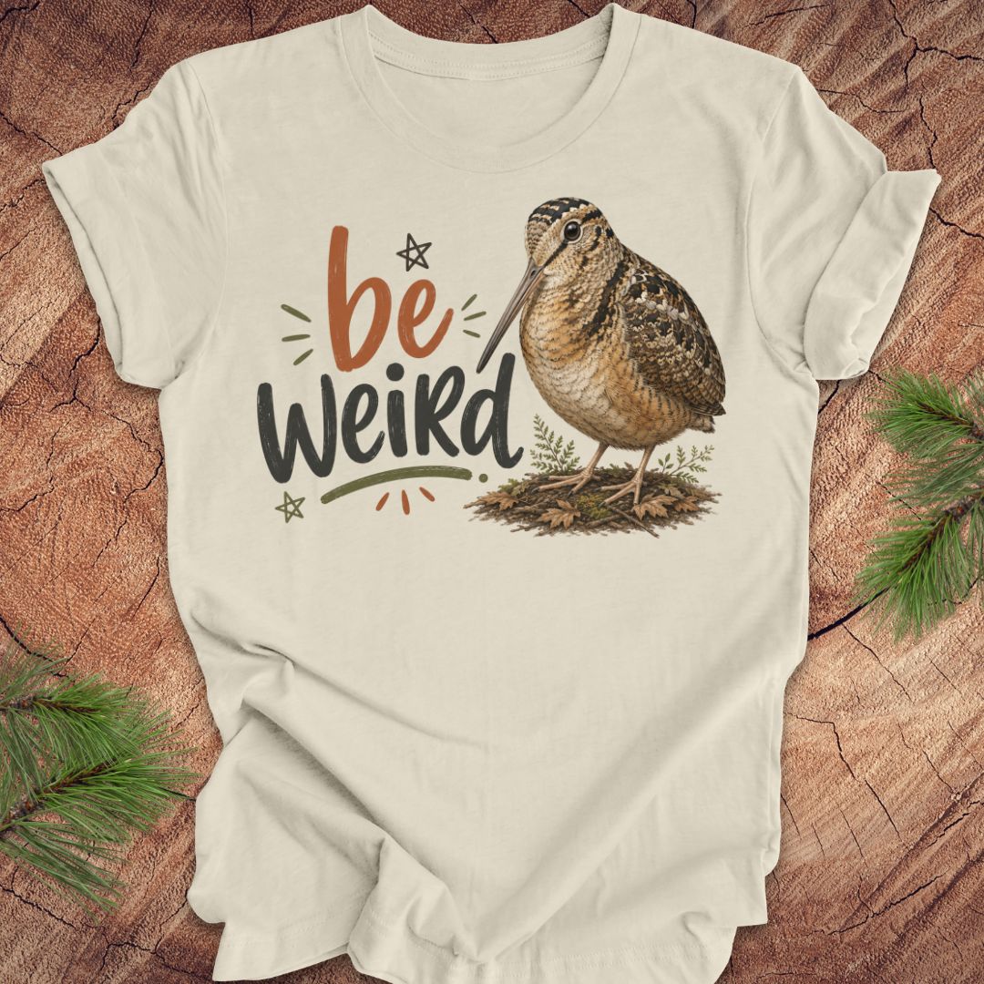 A beige graphic t-shirt featuring the text 'be Weird' in orange and dark gray, alongside an illustration of an american woodcock bird. The t-shirt is displayed flat on a rustic wood background decorated with pine branches.